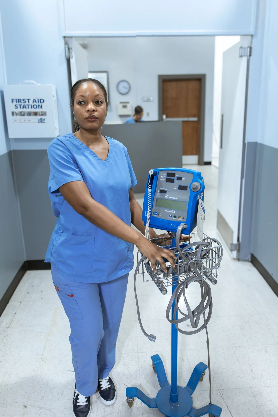 A nurse in scrubs with a vital signs monitor in a clinic hallway
